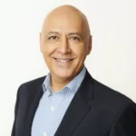 A bald man in a dark suit and light blue striped shirt is smiling at the camera. He has a friendly expression and is standing against a plain white background.