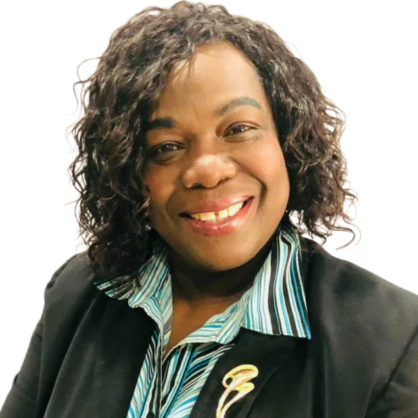 A person with curly dark hair smiles while wearing a black blazer over a blue-striped shirt. The person is also wearing a large brooch on the blazer's lapel. The background is plain white.