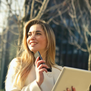A woman with long blonde hair, wearing a white coat, stands outdoors, smiling and looking to her left. She holds a smartphone in her right hand and a tablet in her left. The backdrop of barren trees and a blurred building subtly reflects the essence of Employee Value Proposition in an ever-changing environment.