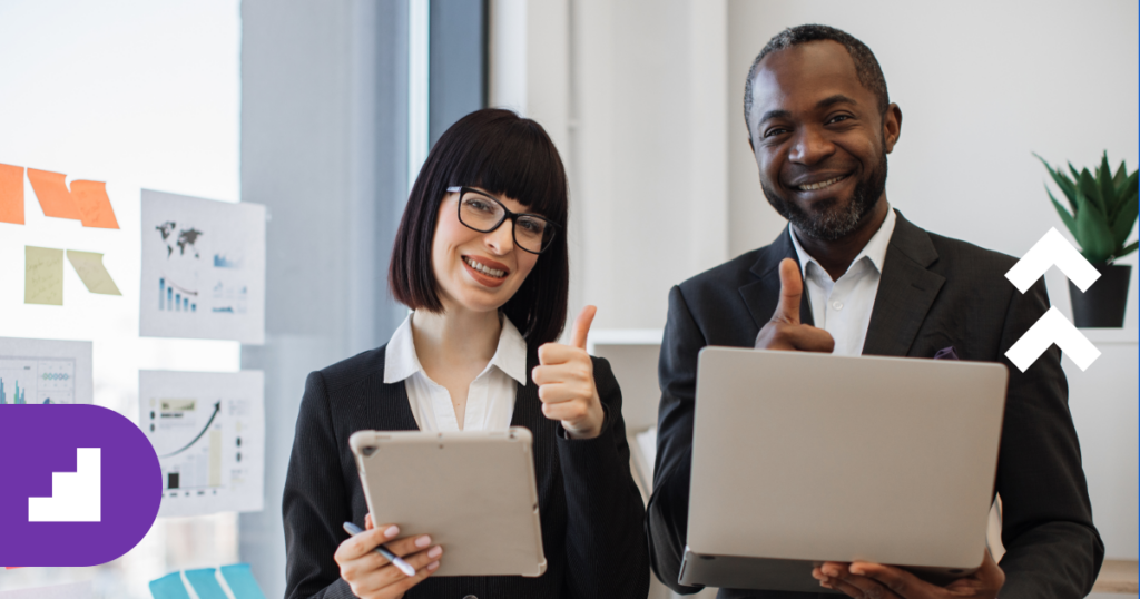 Two business professionals, one woman and one man, stand smiling in a modern office. Both are dressed in suits and give a thumbs-up gesture. The woman holds a tablet displaying the year in review while the man holds a laptop. A wall with charts and graphs is seen in the background.